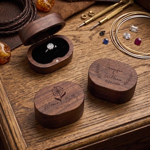 May include: Three oval dark wood ring boxes with magnetic closures. One box is open, revealing a silver diamond ring. The other two boxes are closed and engraved with names, dates, and floral designs: Alexis & Billy 06.06.2024 and Jennifer & William 11.26.2024.