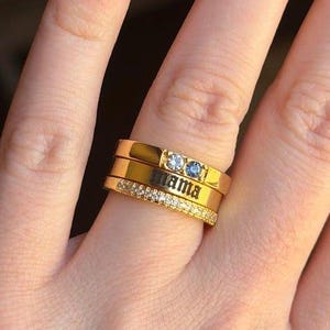 May include: A stack of three gold-toned rings worn on a finger. The top ring features two small blue stones, the middle ring is engraved with the word mama in a black font, and the bottom ring is encrusted with a row of small clear stones.