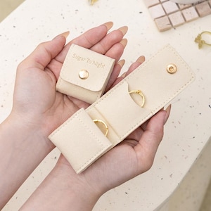 May include: A beige, soft-textured jewelry organizer case featuring a small square pouch with the text Sugar To Night and a gold snap button, alongside a longer rectangular section with two internal slots holding gold rings and a gold snap closure.