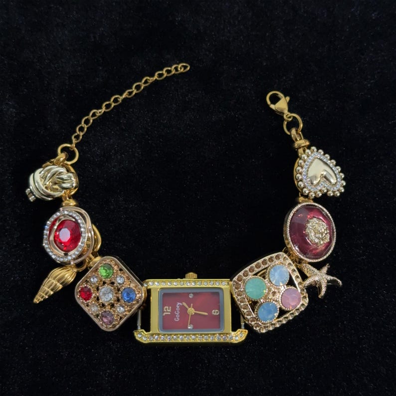 May include: Gold-toned bracelet watch with a rectangular red face and a gold bezel. The bracelet features various charms, including a heart, a seashell, and a starfish, adorned with colorful gemstones.