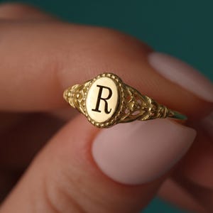 Puede incluir: Un anillo de oro con una cara de forma ovalada con la letra R grabada en el centro. La banda del anillo muestra un intrincado trabajo de filigrana metálica con un borde de cuentas que rodea la placa ovalada central, con un acabado pulido.