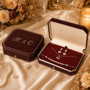 Könnte beinhalten: Eine dunkelrote Schmuckschatulle aus Samt mit dem goldenen Schriftzug M & O auf dem Deckel, die eine goldene Halskette mit rotem Edelsteinanhänger, passende Ohrringe und ein goldenes Armband mit Blattmuster und kleinen weißen Perlen enthält.