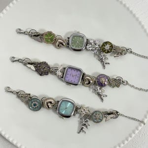 May include: Three silver charm watches with square faces and pastel dials. Each watch has decorative charms, including flowers, hearts, and cherubs. The watch bands have a delicate chain design.
