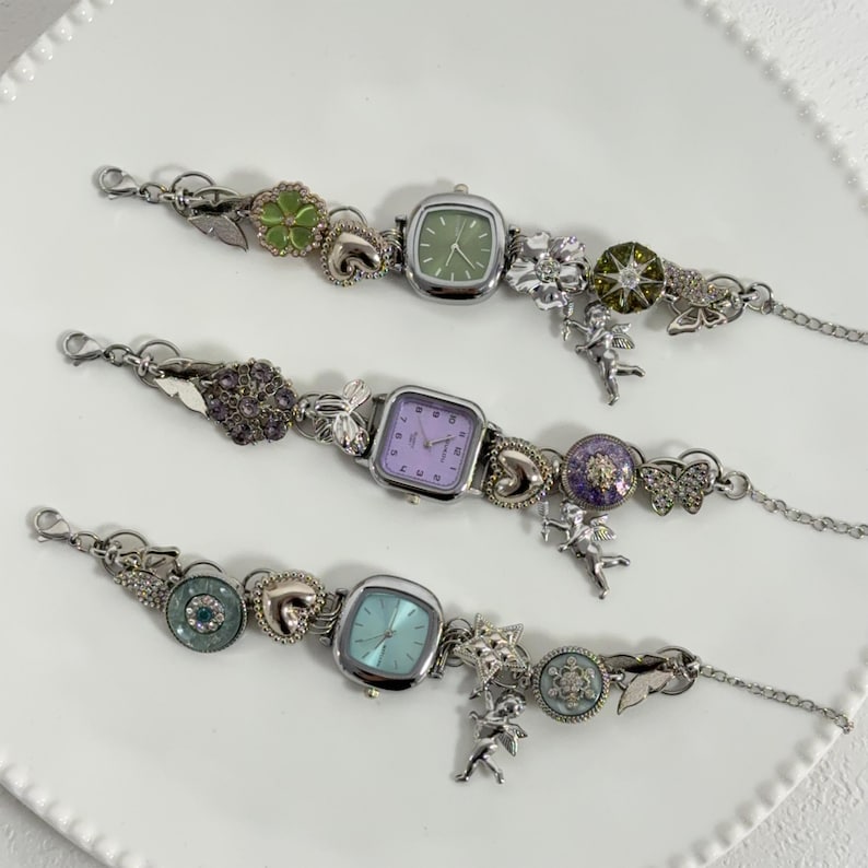 May include: Three silver-toned charm watches with square faces and various pastel-colored dials. Each watch features decorative charms, including flowers, hearts, and cherubs. The watch bands have a delicate chain design.