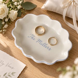May include: An oval ceramic ring dish with a scalloped edge and a light blue rim. The center features the text The Millers in blue script. Two gold-toned metal bands rest inside the dish on the white surface.