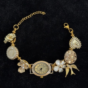 May include: Gold-toned bracelet watch with an oval face and decorative charms. The charms include a knot, a flower, a heart, and a bird. The watch face has gold numerals and hands, and the band is a delicate chain.