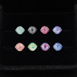 May include: Eight iridescent, square-shaped gemstones with a spiral design are displayed in a black jewelry box. The gemstones are in various colors, including blue, pink, and green. The gemstones are likely for jewelry making.