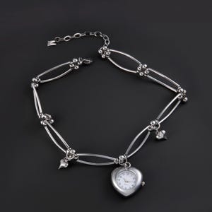 Può includere: Una collana argentata con un pendente a forma di cuore. La collana presenta una serie di maglie ovali, piccoli accenti di perline e ciondoli a forma di cuore. L'orologio ha un quadrante bianco con numeri e lancette nere.