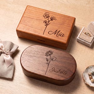 May include: A rectangular light wood box engraved with a flower and the name Mia, and an oval dark wood box engraved with a flower and the name Isabelle. A beige fabric pouch, a small ring box with a gold ring, and a dish with beads are present.