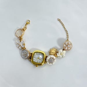 May include: Gold-toned watch bracelet with a square watch face and decorative charms. Charms include flowers, a sun, and a gemstone. The watch face has a white background with gold hands and markers.