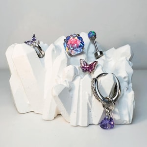 May include: A collection of body jewellery including a silver butterfly hoop earring with purple stones, a large faceted iridescent crystal stud, a small purple butterfly stud, a barbell with a blue opal-like sphere, and a silver hoop with a heart charm.