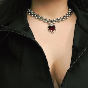 May include: A silver-toned choker necklace with a red heart pendant. The heart pendant has a rose at the top and is surrounded by spikes. The necklace is made of small, interconnected beads and floral-shaped links.