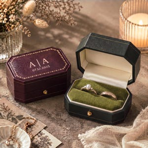 May include: Two octagonal ring boxes with gold-tone metal latches. One box is deep burgundy with gold-stamped text A | A and 07.12.2025. The other is dark green with a cream interior and olive green velvet ring slots holding a pearl ring and a band.