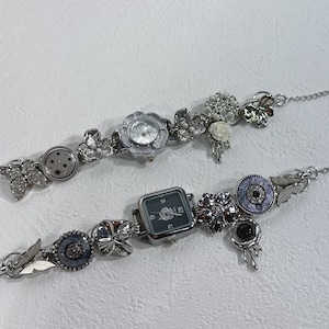 May include: Two silver-toned watch bracelets with floral and decorative accents. One watch has a round face, the other a square face. Both feature intricate details, including flowers, butterflies, and other embellishments. The bracelets are displayed on a white surface.