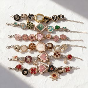 May include: A collection of seven decorative wristwatches with various embellishments. Each watch features a unique design, including floral, heart, and geometric shapes, in colors like gold, pink, and red. The watch bands are silver-toned with chain links.