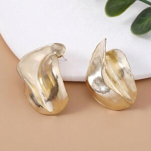 May include: A pair of gold-toned earrings featuring an abstract, organic, and folded metal design. The surface of the earrings has a textured, matte finish with smooth, curved edges that create a sense of depth and movement in the metal structure.