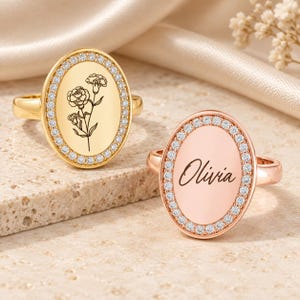 Op de afbeelding: Twee ovale ringen met gepolijste banden. De gouden ring heeft een gegraveerd bloemmotief, terwijl de roségouden ring is gegraveerd met de naam Olivia. Beide ringen hebben een centraal ovaal vlak omringd door een rand van kleine, heldere stenen.