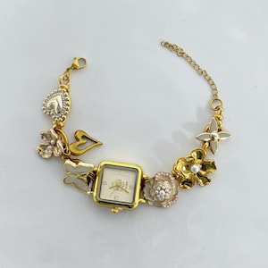 May include: Gold-toned bracelet watch with a square face and a cream-colored dial. The bracelet features decorative charms, including flowers, butterflies, and a heart. The watch has a chain closure.