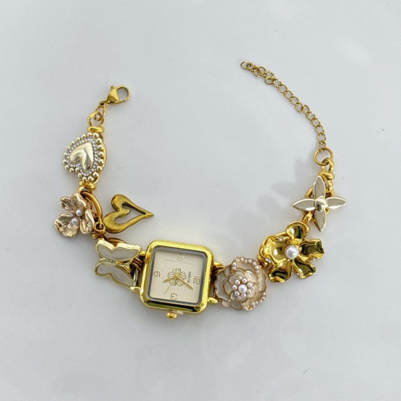 May include: Gold-toned bracelet watch with a square face and a cream-colored dial. The bracelet features decorative charms, including flowers, butterflies, and a heart. The watch has a chain closure.