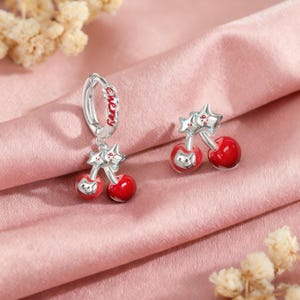 Puede incluir: Pendientes de cereza plateados con cerezas de esmalte rojo y detalles de estrella. Un pendiente tiene la palabra "cherry" en rojo. Los pendientes se muestran sobre una superficie de satén rosa, mostrando un diseño divertido y caprichoso.