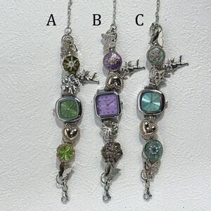 May include: Three silver-toned charm bracelets with square-faced watches. Each bracelet features decorative charms, including hearts, butterflies, and floral accents. The watch faces are green, purple, and teal.