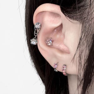 May include: A close-up of an ear adorned with multiple pieces of jewelry. A silver safety pin earring with a dangling paw print charm, a small pink and clear gem paw print stud, and a pink hoop earring with pink gems.