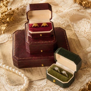 May include: A stack of three octagonal dark red jewelry boxes with gold-tone trim and a single dark green octagonal jewelry box. The open boxes reveal velvet interiors holding two gold flower-shaped rings and two silver-tone diamond rings.