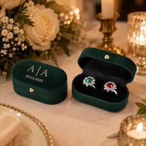 May include: An oval dark green velvet ring box with a gold-colored metal clasp. The lid features the gold-printed text A | A and 07.12.2025. The open box interior holds two silver rings, one with a green stone and one with a red stone.