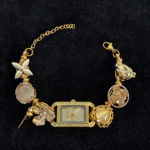 May include: Gold-toned bracelet watch with a rectangular face and a gold band. The watch features a gold bezel with clear stones, a gold dial, and decorative charms, including a flower, a heart-shaped stone, and a knot.