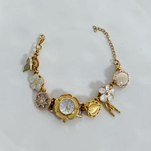 May include: Gold-toned bracelet watch with a floral design. The watch features a white watch face and is adorned with charms, including flowers, a butterfly, and a heart-shaped charm. The bracelet is on a white surface.