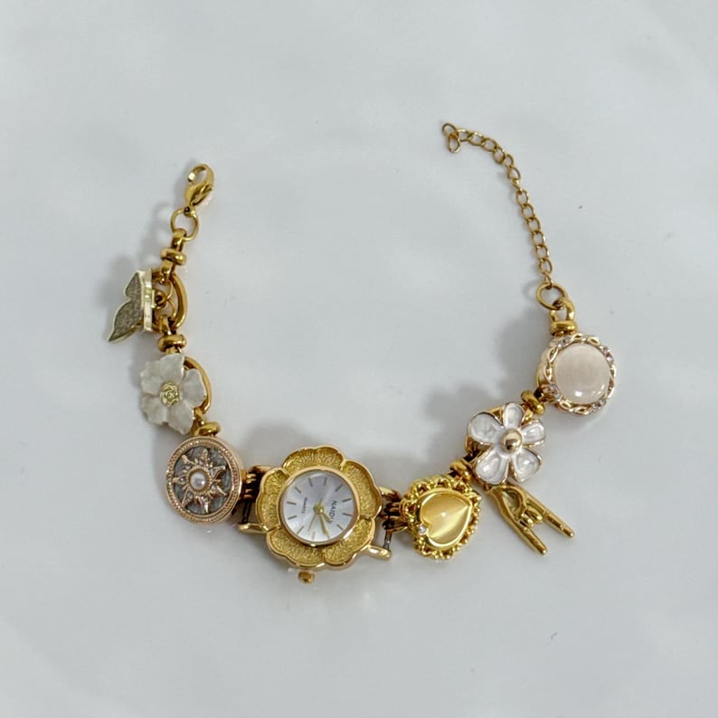 May include: Gold-toned bracelet watch with a floral design. The watch features a white watch face and is adorned with charms, including flowers, a butterfly, and a heart-shaped charm. The bracelet is on a white surface.