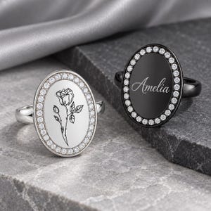 May include: Two oval rings with halo settings of small clear stones. The silver-toned ring features a black line drawing of a rose. The black-toned ring features the name Amelia engraved in a white script font on the center surface.