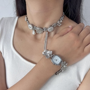 Könnte beinhalten: Silberfarbenes Schmuckset mit Halskette und Armbanduhr. Die Halskette hat dekorative Elemente und eine herabhängende Kette. Die Uhr hat ein quadratisches Zifferblatt und florale Akzente. Das Set enthält Perlen.