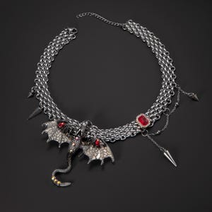Puede incluir: Collar plateado con colgante de dragón, detalles rojos y diseño de cadena de múltiples hilos. Incluye dijes de púas colgantes y una gema roja. Un accesorio de inspiración gótica.