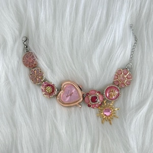 Op de afbeelding: Een roze en goudkleurig armbandhorloge met een hartvormige wijzerplaat. De armband heeft verschillende roze bloemen- en edelsteenachtige bedels, samen met een gouden zon-bedel. Het horloge staat tegen een witte, donzige achtergrond.