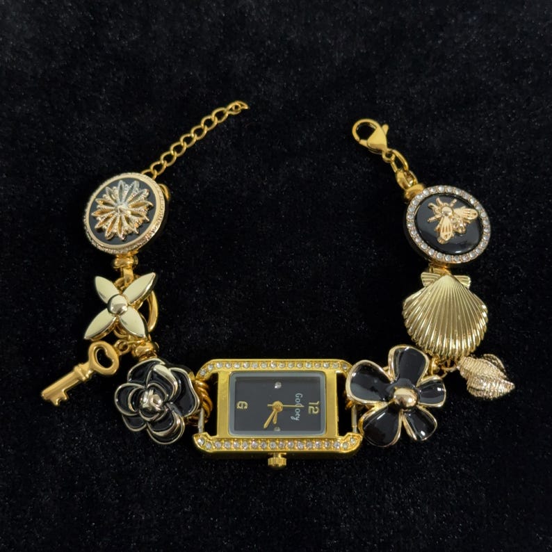 May include: Gold-toned bracelet watch with a black watch face and decorative charms. The watch features a rectangular face with a gold bezel and a black dial. Charms include flowers, a key, a bee, and a seashell.