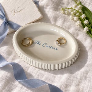 May include: An oval cream-colored ceramic ring dish with a fluted edge. The center of the dish features the blue text 'The Carters' printed in a script font. Two gold-toned rings are placed inside the dish, one plain band and one with a stone.