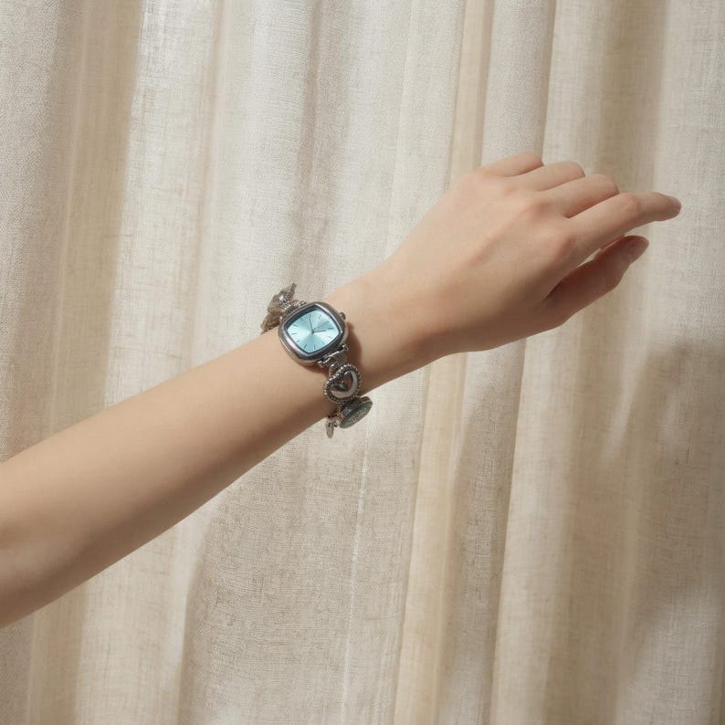 May include: A silver-toned wristwatch with a square, light blue face and a heart-link bracelet. The watch is worn on a wrist, showcasing its design. The watch face has a light blue hue, and the bracelet features heart-shaped links.