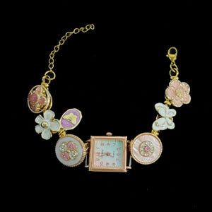 Puede incluir: Un reloj de pulsera dorado con esfera cuadrada y esfera verde claro. La pulsera presenta dijes decorativos que incluyen flores, mariposas y adornos redondos con detalles en rosa y blanco. El reloj tiene un movimiento de cuarzo.