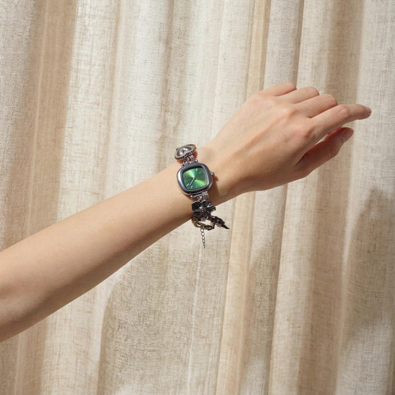 May include: A silver-toned watch with a square green face and a decorative floral band. The watch is on a wrist, showcasing the intricate design and color contrast. The watch band features a unique, ornate design.