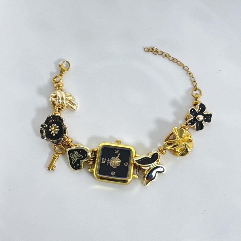 May include: Gold-toned watch bracelet with a square black watch face. The bracelet features black and gold charms, including flowers, butterflies, and a key. The watch has a gold bezel and a black dial with gold numerals.