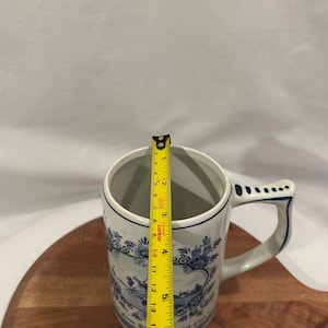 May include: A white ceramic beer stein with blue floral designs and text. The stein is approximately 15 cm tall. The handle and rim are accented with blue. The stein is on a wooden cutting board.