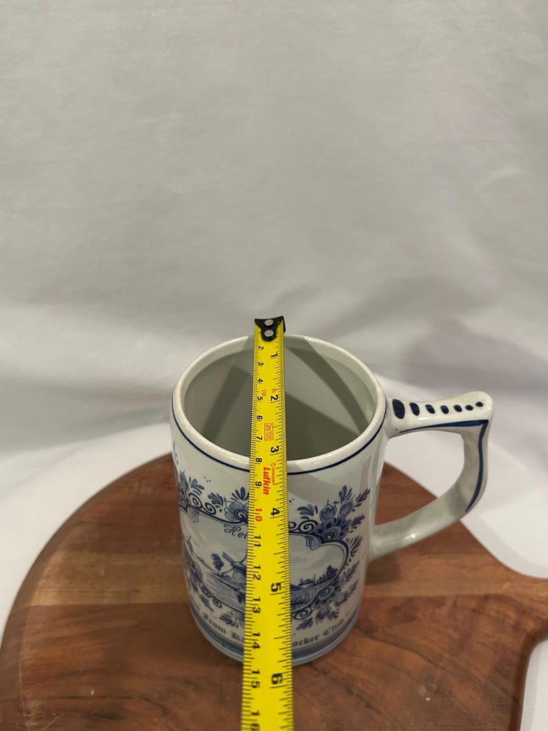 May include: A white ceramic beer stein with blue floral designs and text. The stein is approximately 15 cm tall. The handle and rim are accented with blue. The stein is on a wooden cutting board.