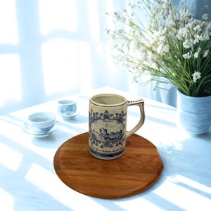 May include: A vintage ceramic beer stein with a blue and white windmill design, a handle, and the text "Heineken." The stein is on a round wooden tray, with two small teacups and a vase of white flowers in the background.