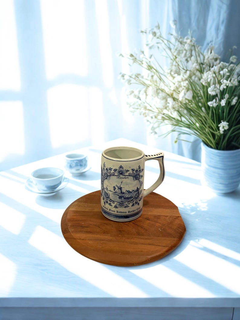 May include: A vintage ceramic beer stein with a blue and white windmill design, a handle, and the text "Heineken." The stein is on a round wooden tray, with two small teacups and a vase of white flowers in the background.