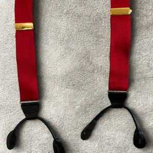 Può includere: Bretelle rosse con chiusure metalliche color oro e attacchi in pelle nera. Le bretelle sono realizzate in un materiale rosso liscio e uniforme e sono progettate per essere indossate con i pantaloni.