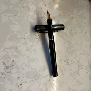 May include: A black fountain pen with a gold nib and gold trim. The pen is lying on its side with the cap next to it. The pen is on a white and gray marble surface.