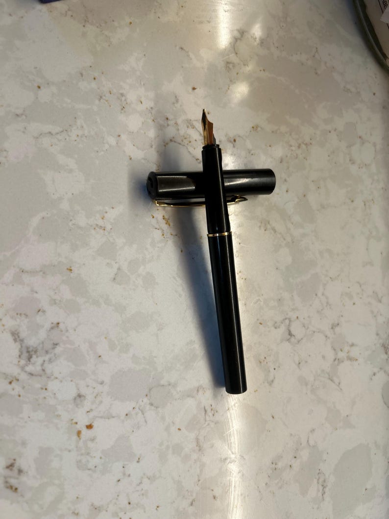 May include: A black fountain pen with a gold nib and gold trim. The pen is lying on its side with the cap next to it. The pen is on a white and gray marble surface.