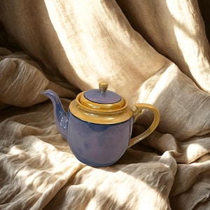 May include: A vintage teapot with a light blue body, a yellow rim, and a matching lid. The teapot has a curved spout and handle, and sits on a beige fabric background. The teapot is a classic design, perfect for serving tea.