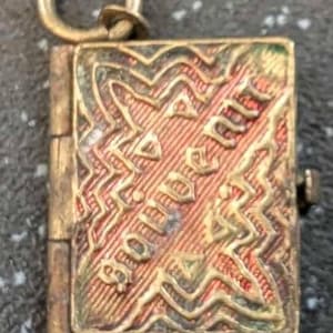 May include: A small, rectangular, hinged pendant with a gold-colored finish. The front features an embossed design with the word "Souvenir" visible. The pendant has a small loop at the top for attaching to a chain or bracelet.